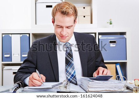 A man writing on a work desk while using a calculator in an office