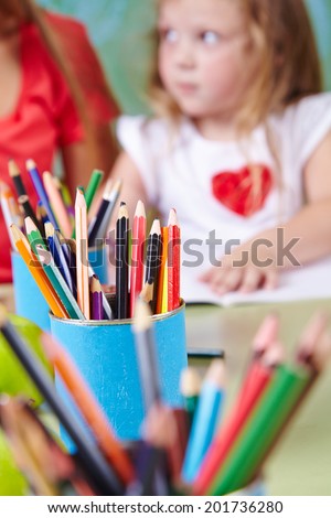 Many crayons on table in kindergarten with a girl drawing in the back