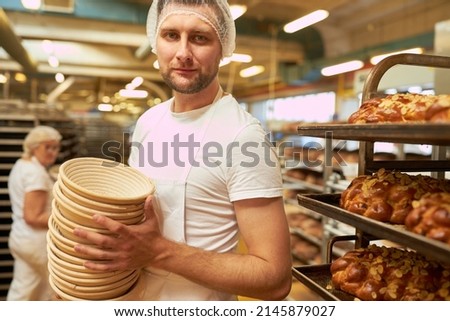 Young man as a baker's apprentice with a stack of bread baskets in front of a shelf trolley with fresh plaited yeast 商業照片 © 