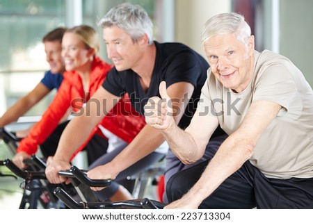 Happy senior man in gym on bike holding his thumbs up