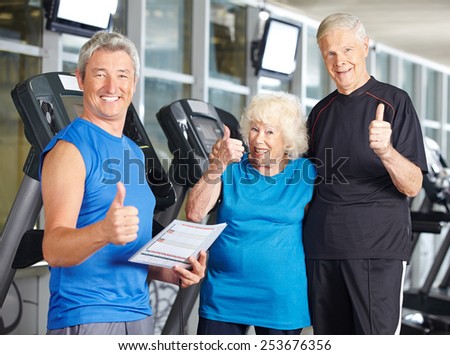 Happy senior couple in gym holding thumbs up with fitness trainer