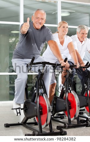 Happy senior man on bike holding thumbs up in gym