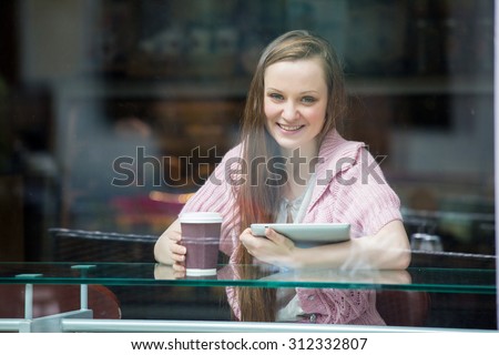 Young pretty woman with long hair drinking coffee and using tablet computer in cafe. Smiling and looking through the window