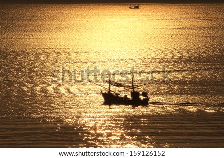 A fishing boat is coming home in the early morning at sunrise from the work of the night in Prachuab, Thailand. It crosses the the golden reflection of the rising sun in the water of the sea.