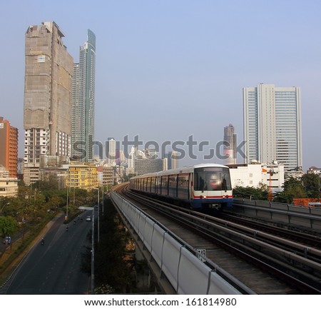 The famous thai skytrain on it\'s elevated track is speeding through the city of Bangkok. In the background are the towers and skyscrapers of the city. Deep down a few cars and mopeds are visible.