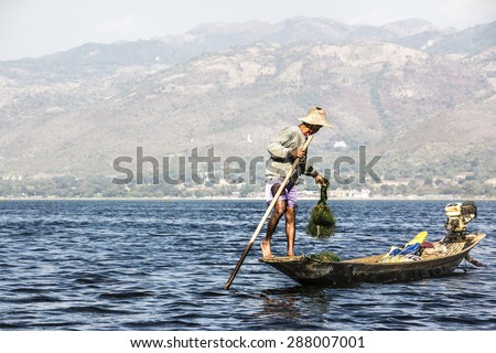 NIAUNGSHWE, MYANMAR - FEBRUARY 2, 2015: A fisherman at lake Inle, Myanmar stands in his boat, rows with one leg, and handles the net. In the background are the mountains.