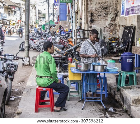MALAMYINE, MYANMR - FEBRUARY 14, 2015: A street seller is cooking food in the main street of Mawlamyine, Myanmar. A customer is sitting and waiting.