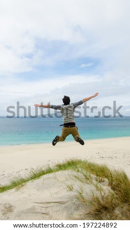 Young model jumping on a sand dune with open arms. White sandy beach and blue sky in the background. Scotland, UK.