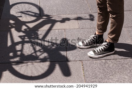 Focus on girl legs wearing black sneakers and skinny brown trousers in front of the shadow of a bicycle projected on the pavement.