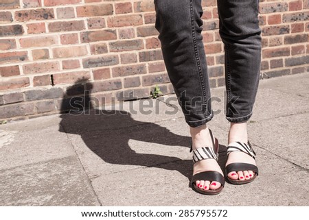 Model wearing black and white sandals with zebra print. Red nail varnish on toes and skinny black jeans. Focus on woman legs and feet. Shadow and brick wall in background.