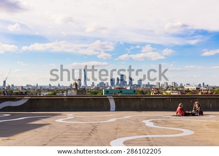Peckham, London, UK - June 06 2015: Two girls sit on the floor of a rooftop bar cafe in south-east London.View of the City of London skyscrapers and the Shard in the background.