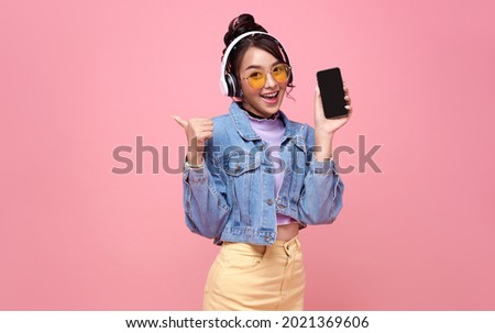 Young Asian teen woman showing smart phone she listening music in headphones isolated on pink background.