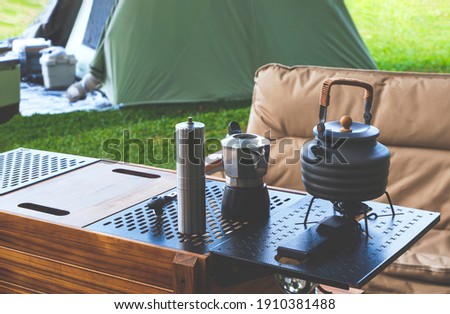 Vintage outdoor black kettle and coffee grinder on camping table with brown armchair on green lawn in camping area