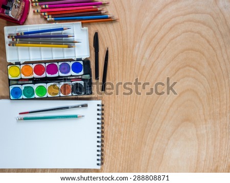 Top view of watercolor paints, color pencils, brushes for painting with blank  sketchbook on  wooden background