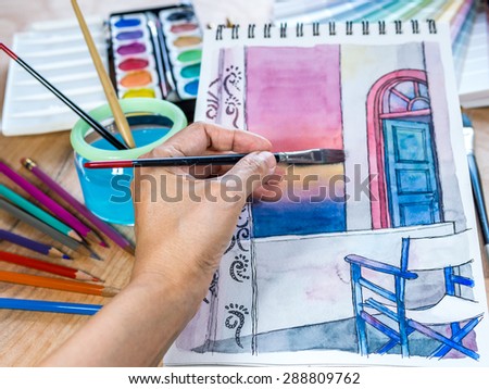 Hands painting architectural on sketchbook with watercolor paints, color-pencils over  wooden table background