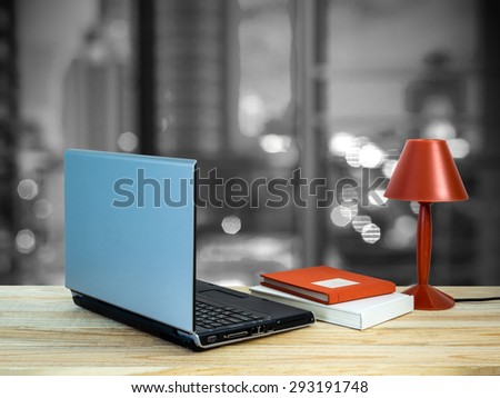 Red lamp, laptop computer, books on wooden table top over bokeh cityscape background/ interior still life