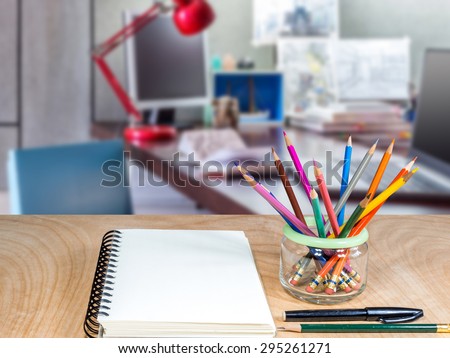 Color pencils in glass jar with notebook on wooden tabletop over  modern office  background