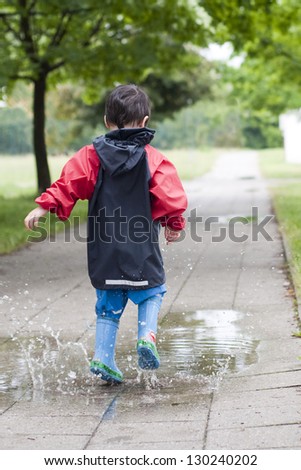 Small boy jumping in a puddle on the path in a park after a rain.