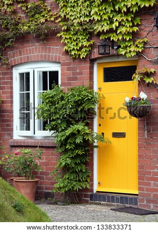 Yellow front door entrance and old style window of a red brick house or a cottage with hanging flower basket and green ivy.