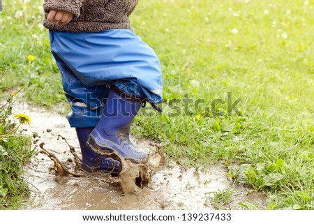 Child in boots jumping in muddy puddle after rain.