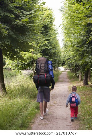 Father and small child with backpacks walking or trekking on a path along a river or canal.