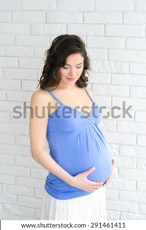 young beautiful pregnant woman in blue shirt holding her belly on white brick wall background