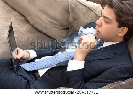 Businessman relaxing on a sofa smoking and working puffing on an electronic cigarette or e-cigarette as he reads his tablet computer