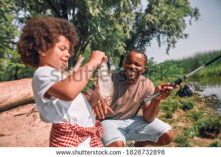 Cute curly Afro-American boy looking at the fish in his hands while his dad holding a fishing rod