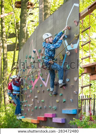 Mother and son in a helmet with safety harness climb the wall on the background of trees and foliage