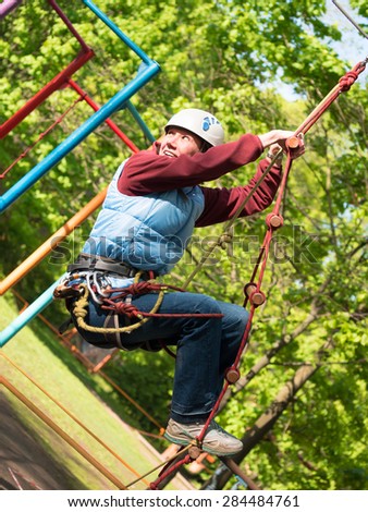 Adult woman in a helmet and with a safety system climbs a rope ladder on the background of trees with leaves
