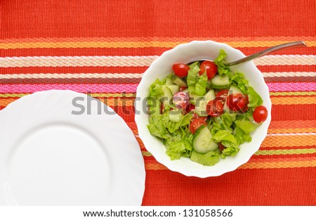 A green salad in a stylish white bowl. With rocket leaves cherry tomatoes spanish onions and capsicum. Empty white plate near.