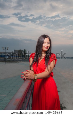 Bright portrait of classy brunette in red shiny dress and long straight hair . Vertical shot against sky, outdoors. Luxury photography.