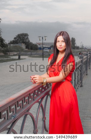 Fashion portrait of classy brunette in red shiny dress and long straight hair . Vertical shot against sky, outdoors.