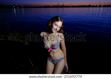 A brunette bikini model with small breasts posing on a beach. Sexy girl in bikini posing at sunset against blue water