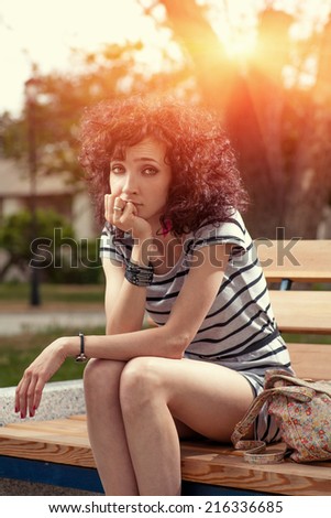 Portrait of young girl sitting on a bench in spring park in sunny day. Redhead girl sitting on the bench in city park