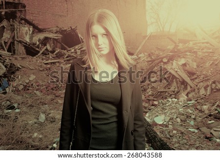 Young blond haired girl posing alone in slums backlit, toned colorized image. Serious calm women in trendy black wear is posing in destroyed shanties area hotspot on her blond hair. Warm toned color.