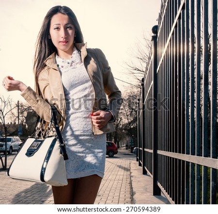 Asian girl is posing with trendy handbag. Stylish beautiful japanese woman in white dress and fashion bag is standing near fence backlit by sunset. Young woman is posing in the street. Instagram color