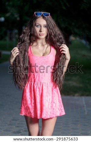 Long haired brunette in pink dress posing in city park at dawn. She is looking very cute.