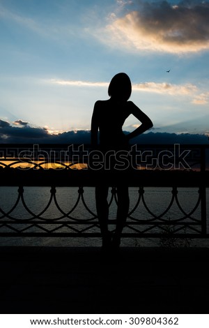 A silhouette of a fashionable woman leaning back against fence and deep colorful evening sky over her.