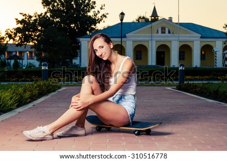Pretty young fashion sexy girl is sitting on skateboard on the park lane in evening summer time have fun and shows her nice bare legs in jeans shorts hipster style photo color filter toned colorized
