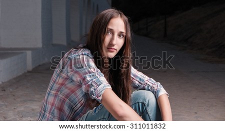 Young cute women is sitting on the ground with her hands on knees. She is looking at camera and wearing checkered shirt and blue slim jeans