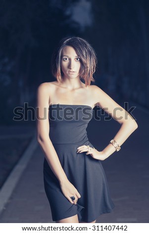 Young latino  women in short black dress is posing outdoors in night time (or late evening) in fashion manner with her hand on waist. She is lit by street lamp from the front side. Alley on background