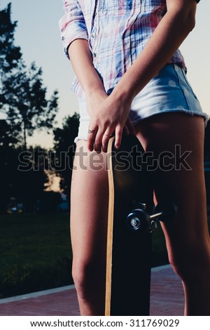 Sporty girl is standing outdoors with her skateboard between her legs with hands crossed on the top of board. Low angle closeup of girl legs with skateboard.  Toned image. Warm color filter