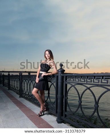 Fashion women leaned back against metal fence with black clutch in her hands in evening time. Sunset sky is over her and a lot of space for text on it. Full body sexy slim girl portrait in black dress