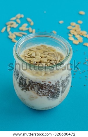 Overnight oats ingredients in a jar on blue background. Mixture of rolled oats, chia seeds, sunflower seeds, yogurt, milk and honey.