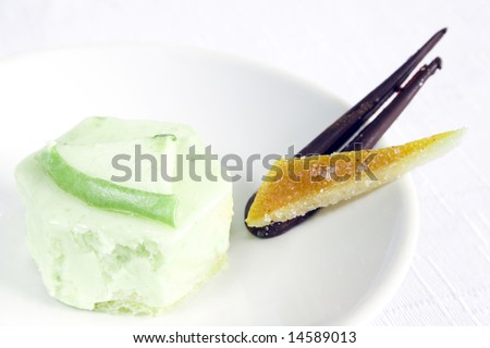 Close up of an apple petit four on a white dish and background. Main focus on the middle part of the PF. It\'s accompanied with a slice of glazed orange skin and a piece of chocolate.