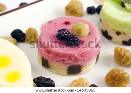 A close up of a red fruits petit four with blueberry and grape on top, surrounded by others petit fours and raisins. Main focus on the central petit four.