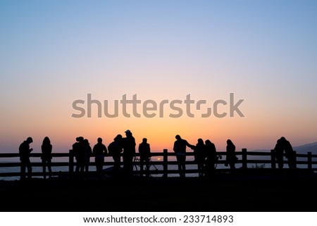 Silhouettes of about fourteen people against a fence, watching the sunset over the ocean with a cloudless sky.