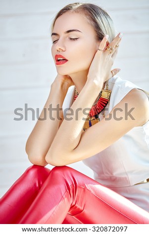 Beautiful fashionable slim girl sitting in red leather pants, a white tank top and gold jewelry.