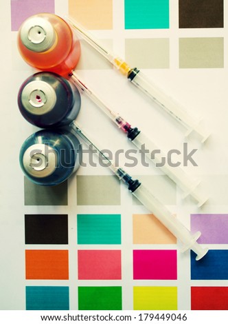 Bottle of ink printer has three colors. Colors is red, yellow and blue with syringe on colored paper.